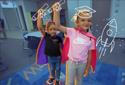 two kindergarten children wearing super hero capes have their hands triumphantly in the air in a classroom